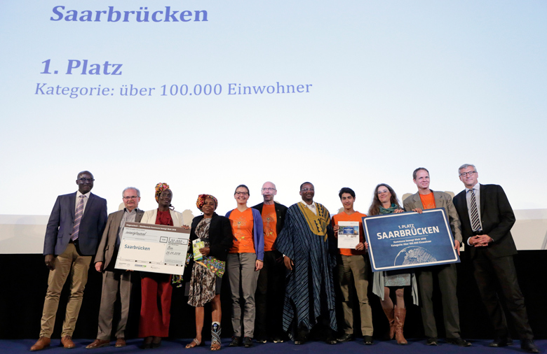 Eine Gruppe, die das Ortsnamensschild Saarbrücken trägt, lächelt in die Kamera.
